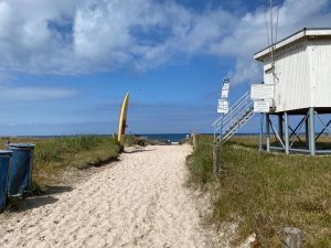 Zu sehen der sandige Weg direkt an die Ostsee im Hintergrund. Links ein Surfboard, rechts die erhöhte Station der DLRG, also der Rettungsschwimmer