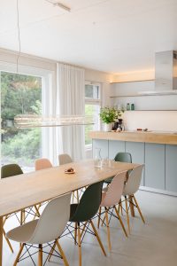 Großzügie Wohn-Essküche mit großer Glasfront und Blick in den Garten. Über dem eleganten Holzisch eine moderne Leuchte. Kochinsel im Hintergrund. Alles hell und freundlich