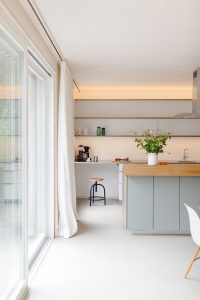 Großzügie Wohn-Essküche mit großer Glasfront und Blick in den Garten. Über dem eleganten Holzisch eine moderne Leuchte. Kochinsel im Hintergrund. Alles hell und freundlich