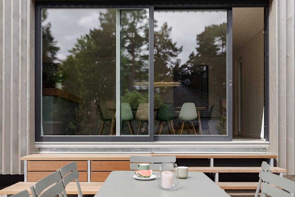 Blick über den grünen Metalltisch auf die Glasfront zu einer Wohnessküche. Innen farbige Stühle und eine moderne Leuchte über dem Holzesstisch