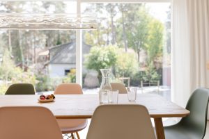 Blick aus der Wohn-Essküche in den Garten. Im Vordergrund Holztisch mit gemütlichen Stühlen und Wassergläsern auf dem Tisch. Moderne Leuchte über dem Tisch