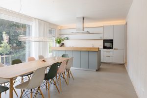 Großzügie Wohn-Essküche mit großer Glasfront und Blick in den Garten. Über dem eleganten Holzisch eine moderne Leuchte. Kochinsel im Hintergrund. Alles hell und freundlich