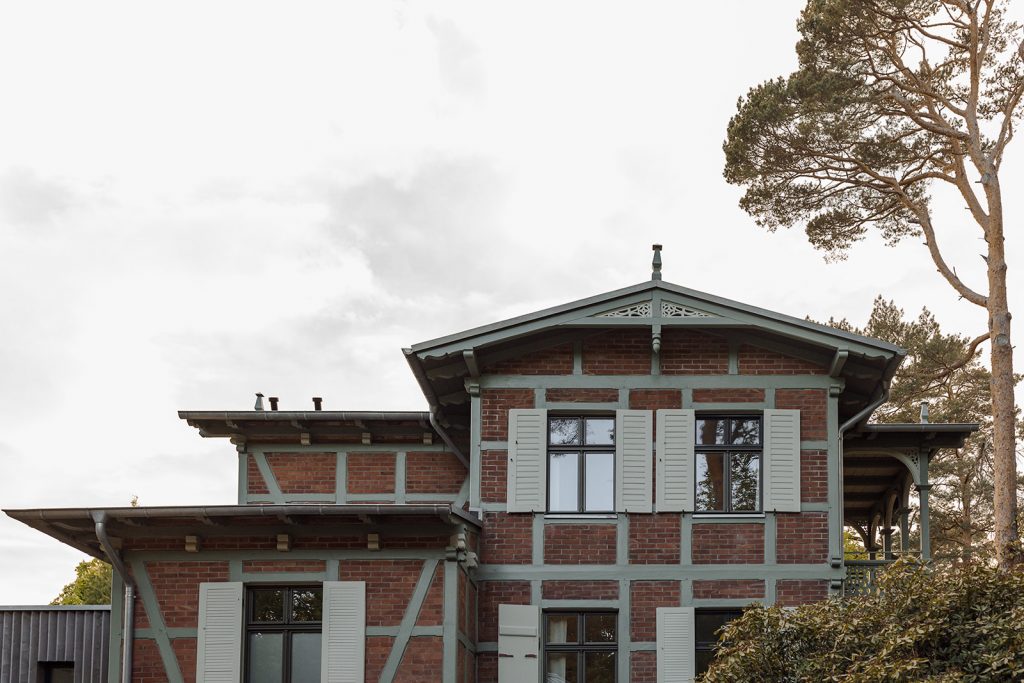 Prächtiges Fachwerkziegelhaus mit alten Schnitzerreien um Dachgibel. Balken sind in einem hellen Olivgrün, die Ziegel klassisch dunkelrot. Rechts ein überdachter Fachwerkbalkon. Links ein moderner Anbau aus Holz.