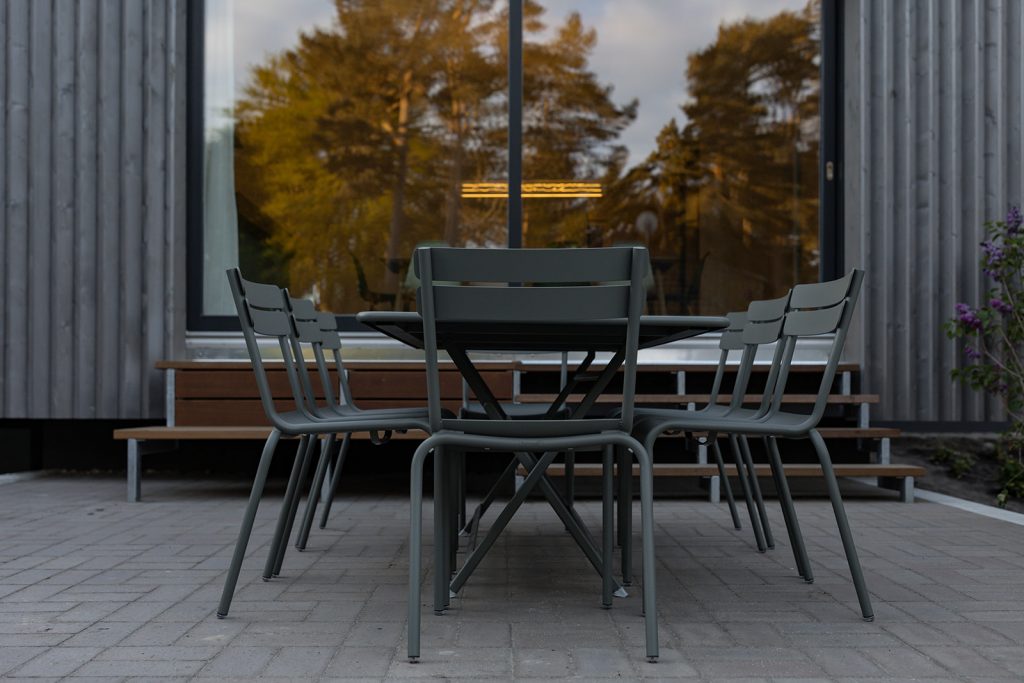 Stühle auf der großen Terrasse. Im Hintergrund spiegeln sich Bäume in der großen Glasfront des Anbaus. Innen Licht. Rechts blühender Flieder