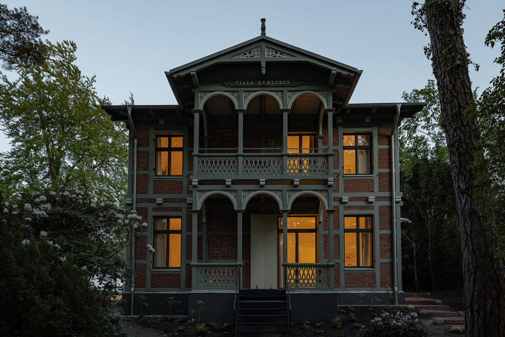Prächtige Fachwerkziegelvilla von Ende 1890. Im Vordergrund zwei überdachte Holzbalkone mit Schnitzerreien. Bild in Abenddämmerung. Inne nhell beleuchtet. Im Vordergrund die Treppe zum Balkon im Erdgeschoss.