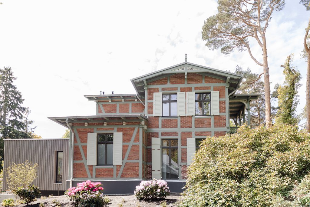 Prächtiges Fachwerkziegelhaus mit alten Schnitzerreien um Dachgibel. Balken sind in einem hellen Olivgrün, die Ziegel klassisch dunkelrot. Rechts ein überdachter Fachwerkbalkon. Links ein moderner Anbau aus Holz. Riesiger Rothodendronbusch vorne rechts. Es scheint die Sonne